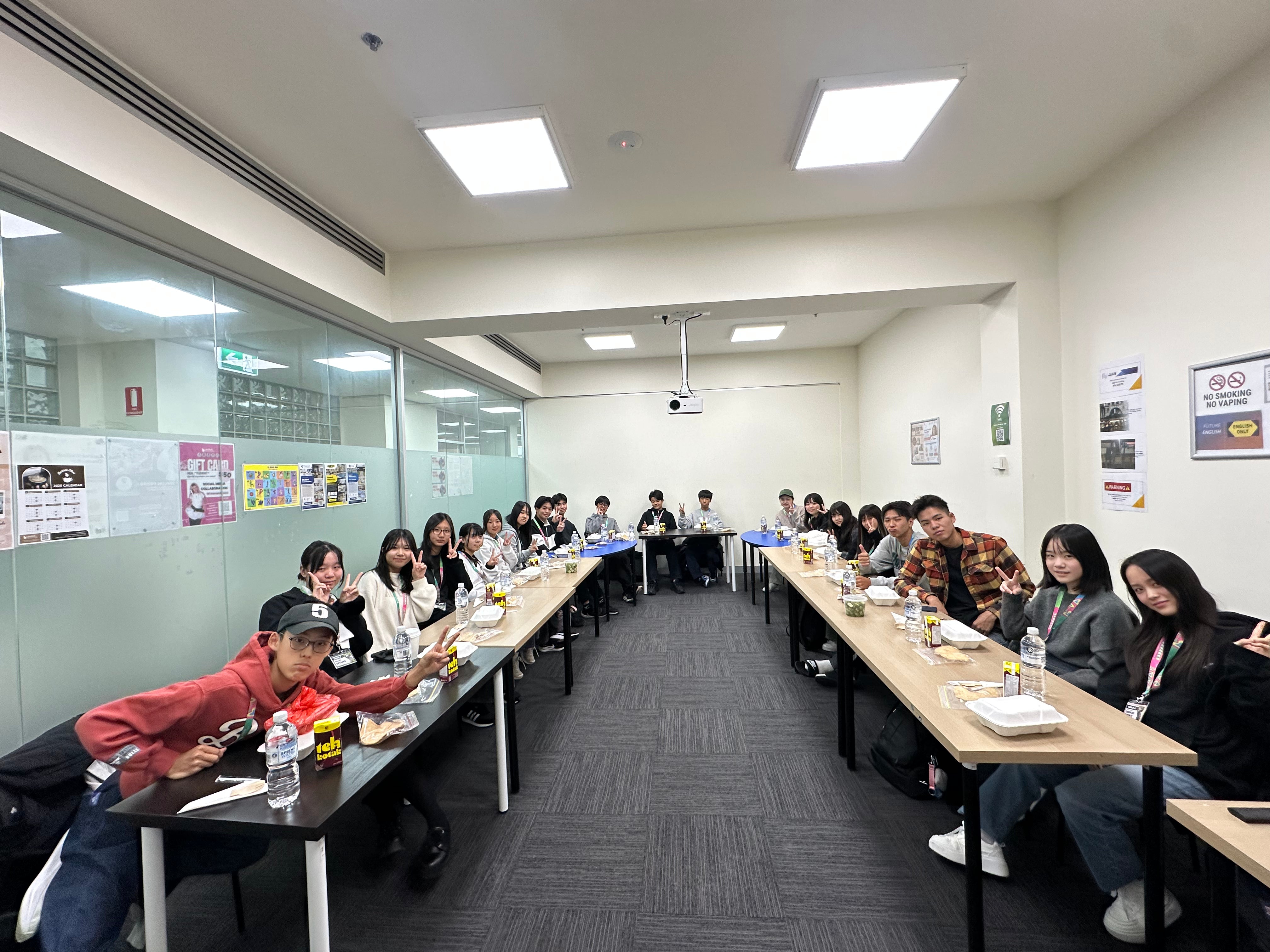 Group of people sitting at long tables in a classroom setting with posters on the wall.
