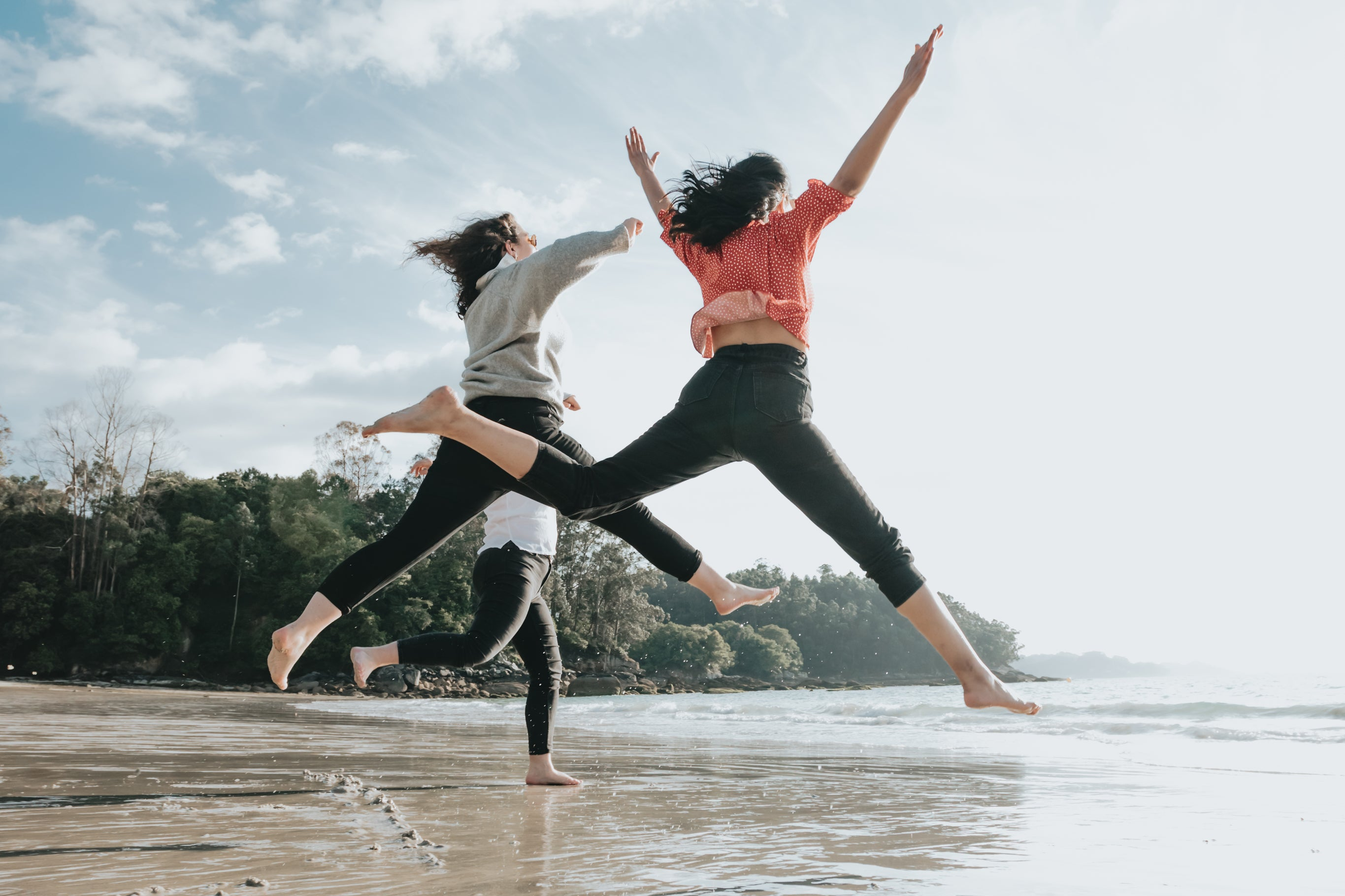 Two people jumping in the air on a beach with trees in the background
